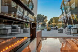 Modern glass-walled lounge with outdoor seating, a row of flame-lit fires, and trees visible through the glass at dusk/dawn light.
