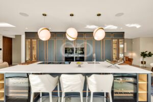 Open-plan kitchen with a white island, three pink globe pendant lights, dark blue cabinets with copper trim, built-in ovens in the center.