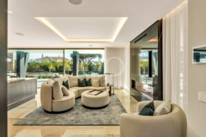 Open-plan living room with beige curved sofa, round ottomans, and large glass walls showing a pool and palm garden outside, with warm ceiling lighting.