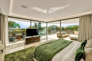 Luxurious hotel bedroom with green bedding and a view of a sunny balcony garden through floor-to-ceiling glass doors