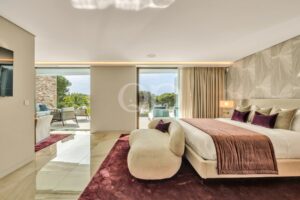 Luxurious hotel bedroom with a king bed, burgundy runner and pillows, cream upholstered furniture, and sliding glass doors to an outdoor seating area.