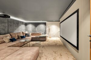 A home theater lounge with a large wall-mounted projector screen on the right, beige sectional sofa, and a ceiling-mounted projector on the left.