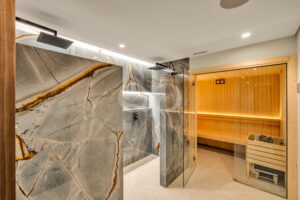 Luxurious shower area with gray marble walls, ceiling rain shower, and a glass door to a wooden sauna room