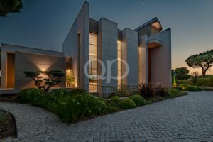 Casa branca moderna com paredes verticais altas e luzes interiores acolhedoras ao anoitecer, num cenário paisagístico bem cuidado.
