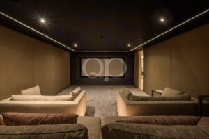 Sala de cinema em casa com iluminação suave, com sofás bege dispostos em forma de U, virados para um grande ecrã situado no fundo da sala.