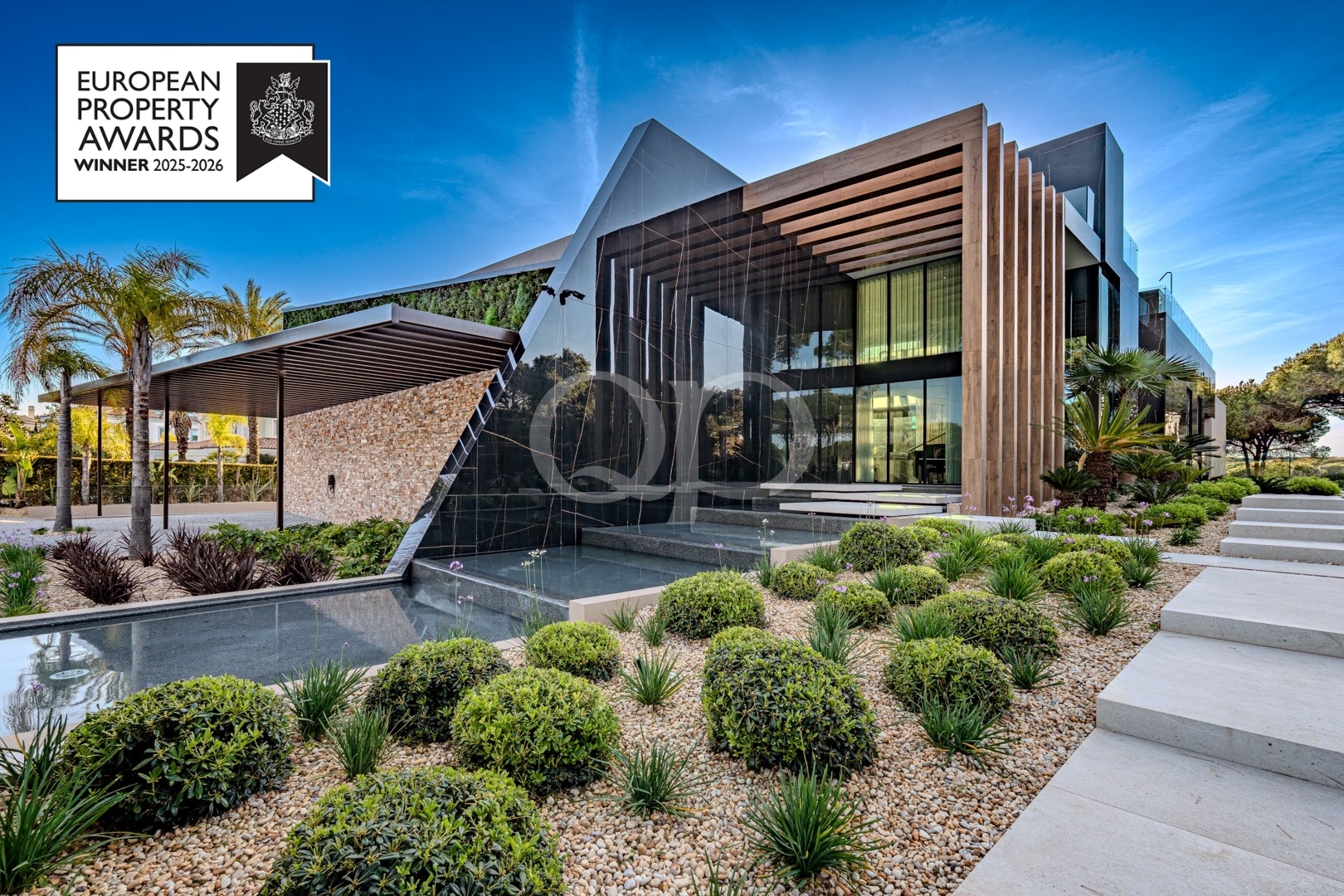 Modern glass-and-stone building with wooden fins, desert landscaping, and a European Property Awards badge in the top-left.