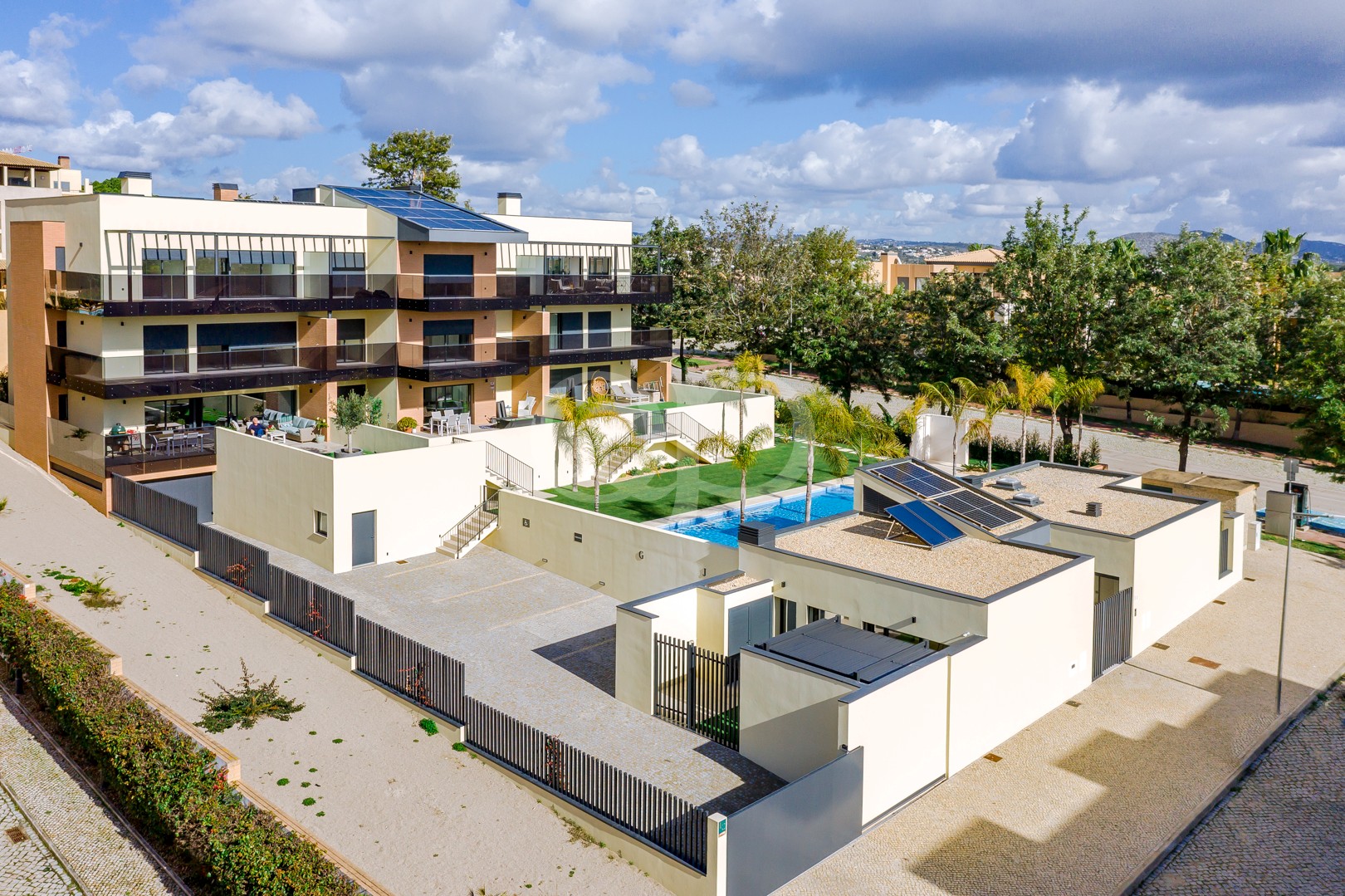 Complexo de apartamentos moderno com várias varandas, painéis solares no telhado e um pátio central com uma piscina azul e palmeiras sob um céu parcialmente nublado