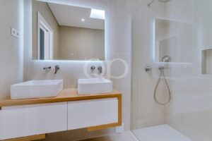 Casa de banho moderna com dois lavatórios retangulares sobre um móvel de madeira, espelho retroiluminado e cabina de duche com divisórias de vidro, revestida com azulejos bege.