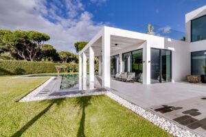 Une maison blanche moderne dotée d'une terrasse couverte, de baies vitrées et d'une longue piscine rectangulaire dans laquelle se reflète le ciel bleu.