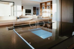 Cozinha e sala de estar em plano aberto, com uma divisória de vidro ao longo de uma ilha de madeira; um prato branco repousa sobre o balcão, e, ao fundo, avistam-se prateleiras embutidas e um televisor.