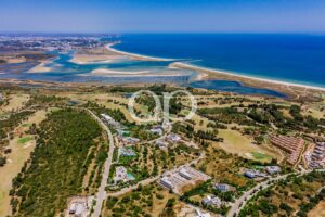 Vista aérea de uma paisagem costeira com o mar azul, praias de areia e uma zona residencial verdejante no interior.
