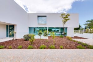 Front view of a modern white house with large glass windows and a landscaped, mulched yard under a blue sky with clouds.