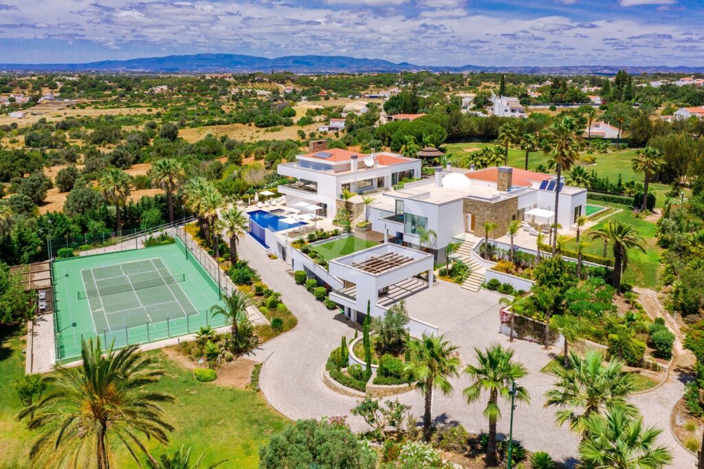 Vista aérea de uma moradia moderna e luxuosa com uma piscina retangular, um campo de ténis e jardins ladeados por palmeiras.