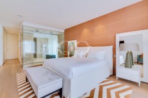Cama king-size com roupa de cama branca num quarto de hotel luminoso e moderno, com uma parede de destaque em laranja e uma casa de banho com paredes de vidro. I