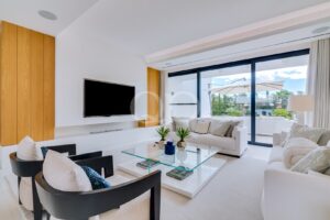 Sala de estar luminosa e moderna, com sofás brancos, uma mesa de centro de vidro e uma televisão fixada na parede, que dá acesso a um pátio através de grandes portas de correr.