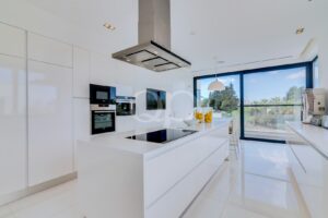 Cozinha branca, luminosa e moderna, com uma ilha, fornos embutidos e um exaustor em aço inoxidável, com vista para uma ampla janela que dá para a vegetação no exterior.