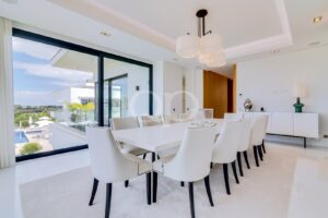 Sala de jantar moderna e luminosa, com uma longa mesa branca e cadeiras estofadas brancas, e grandes portas de vidro que dão acesso a uma piscina exterior.
