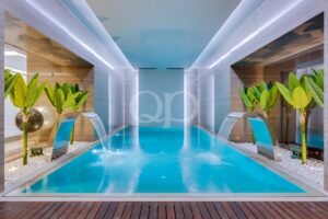 Piscina interior simétrica com dois arcos metálicos curvos de onde jorra água para uma piscina de um azul brilhante, ladeada por plantas tropicais.