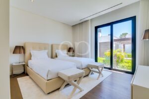 Quarto de hotel com duas camas individuais, roupa de cama branca e cabeceiras bege, junto a portas de correr de vidro que dão acesso a um jardim ensolarado.