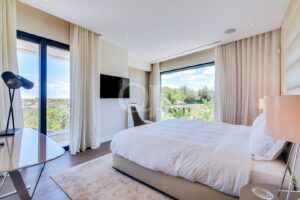 Quarto de hotel moderno e luminoso, com janelas amplas e vista para a vegetação; roupa de cama branca e detalhes em bege.