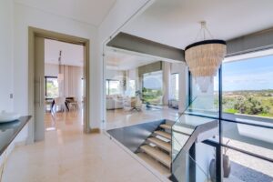Átrio luminoso e moderno, com uma escadaria com corrimão de vidro com vista para uma sala de estar bem iluminada e grandes janelas que revelam a vegetação no exterior.