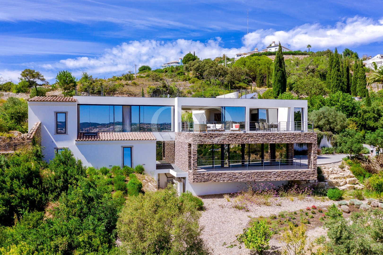 Villa moderne à flanc de colline, avec de grandes baies vitrées, un crépi blanc et une terrasse en pierre, nichée dans un écrin de verdure sous un ciel bleu.