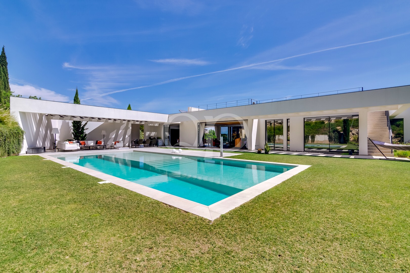 Villa moderne dotée d'une piscine rectangulaire turquoise, d'une terrasse blanche et d'une vaste pelouse sous un ciel d'un bleu éclatant.