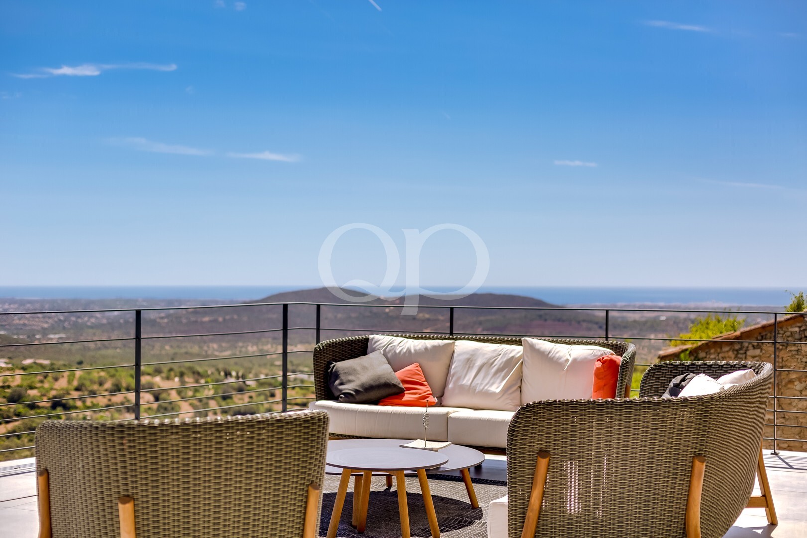 Terrasse extérieure aménagée avec un canapé et des chaises en osier, vue sur le ciel bleu et la mer au loin depuis le flanc d'une colline
