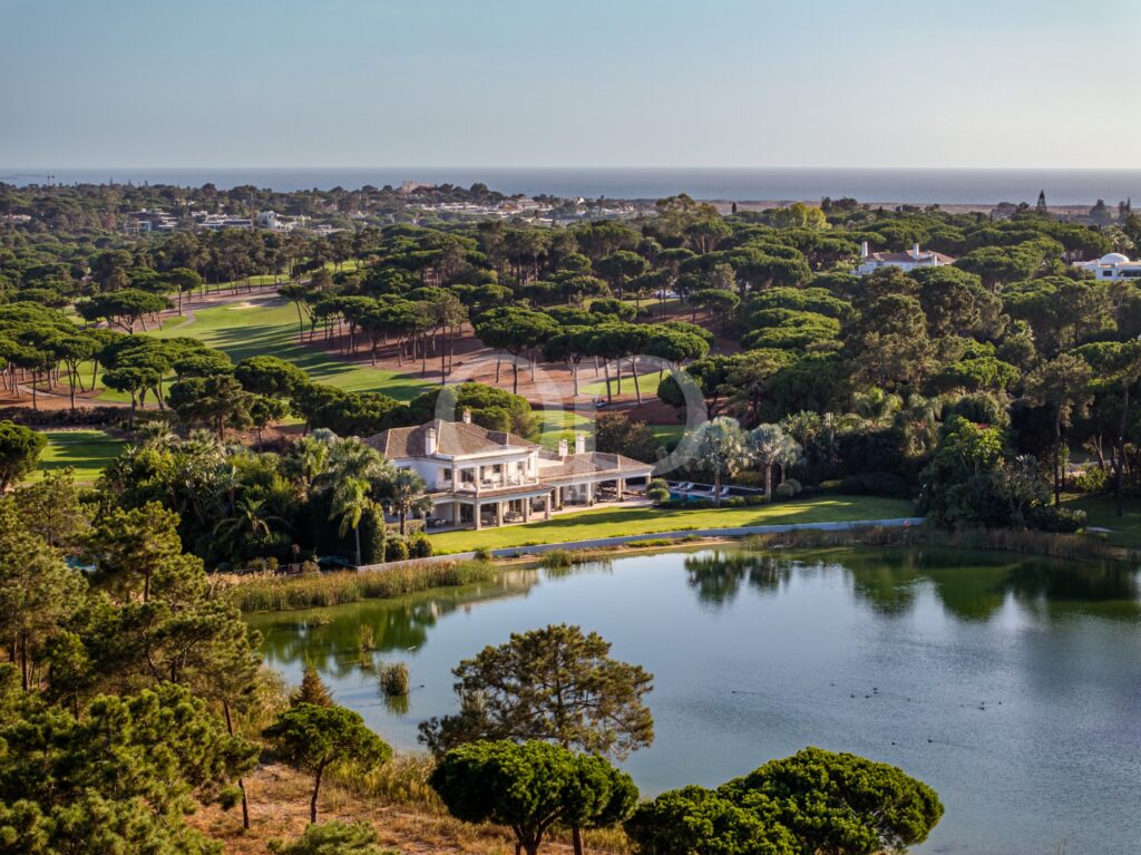 Luftaufnahme eines Golfplatzes an der Küste mit grünen Fairways, dichten Bäumen und einem ruhigen Wasserlauf im Vordergrund, im Hintergrund das Meer.