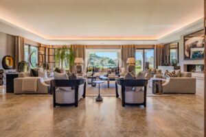 Ein geräumiges, modernes Wohnzimmer mit beigen Sofas, Kissen mit Zebramuster, runden Couchtischen und großen Fenstern mit Blick auf einen sonnigen Garten.