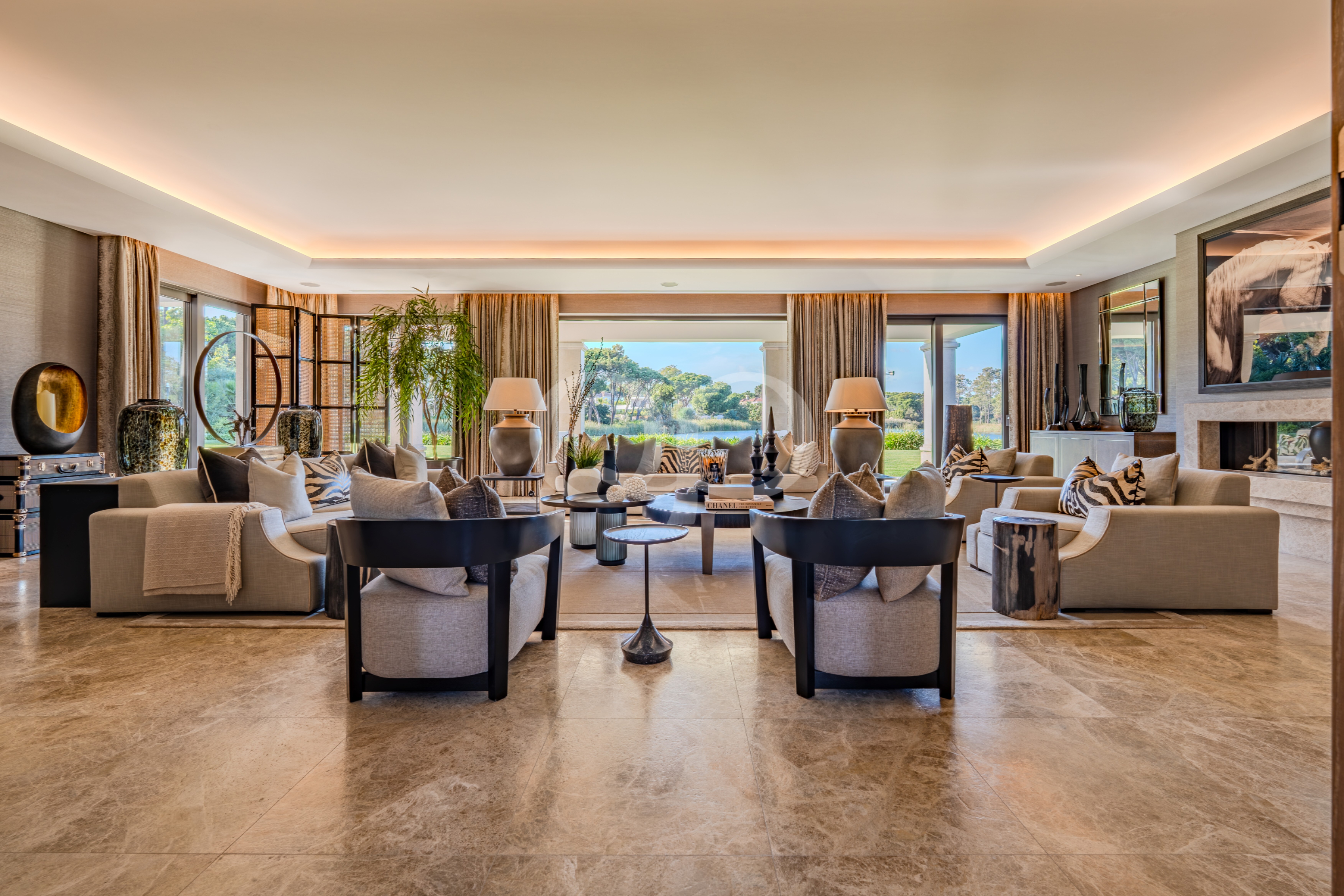 Ein geräumiges, modernes Wohnzimmer mit beigen Sofas, Kissen mit Zebramuster, runden Couchtischen und großen Fenstern mit Blick auf einen sonnigen Garten.
