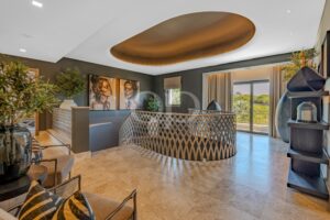 Moderne Hotellobby mit einer großen ovalen Deckenkonstruktion aus Bronze, dunklen Wänden, einem Gittergeländer und einer Sitzecke an einer Fensterwand mit Pflanzen und Kunstwerken.