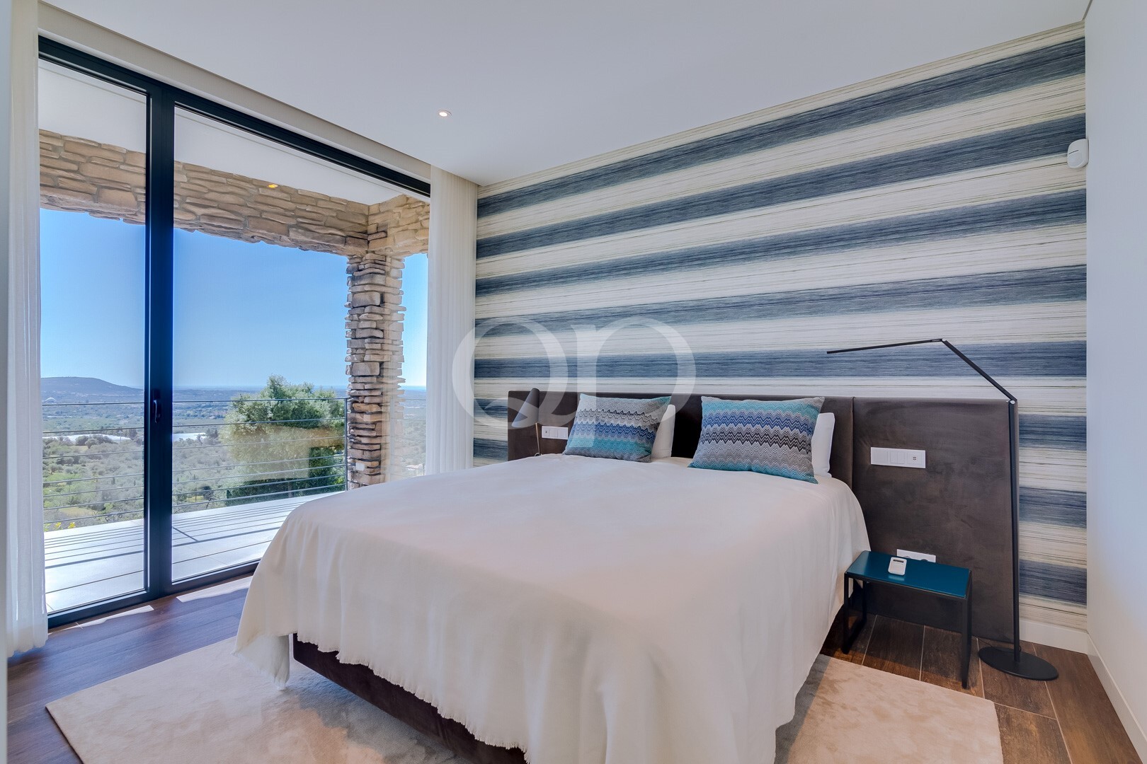 Chambre lumineuse et moderne, avec un lit blanc et un mur d'accent à rayures bleues ; de grandes portes vitrées s'ouvrant sur un balcon en pierre et offrant une vue sur le paysage.