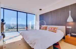 Chambre d'hôtel lumineuse et moderne, dotée de baies vitrées donnant sur les collines ; lit king size avec couette blanche et coussins hexagonaux colorés.