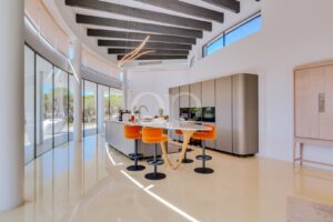 Bright open-concept kitchen with a curved glass wall, central island and orange stools under dark ceiling beams.
