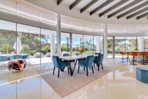 Bright contemporary dining area with glass walls, blue upholstered chairs around a wooden table and a floral centerpiece on a light rug.