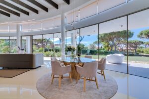 Bright open-plan dining area with a round glass table and beige chairs, large glass walls overlook a landscaped garden.