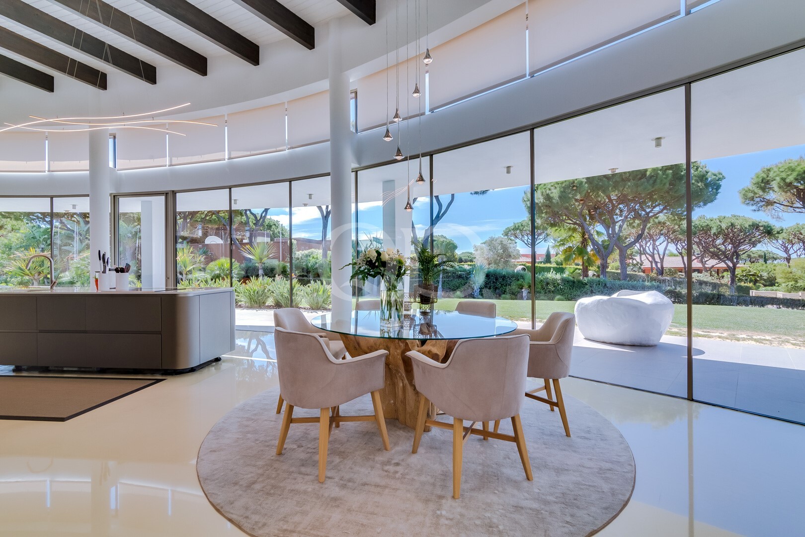 Bright open-plan dining area with a round glass table and beige chairs, large glass walls overlook a landscaped garden.