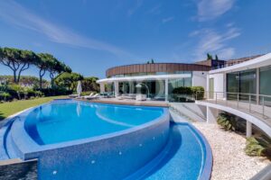 Luxurious modern villa with a curved blue swimming pool and outdoor lounge area under a clear sky, surrounded by trees and greenery.