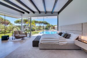 Luxurious bedroom with floor-to-ceiling glass walls overlooking a pool and lush garden outside.
