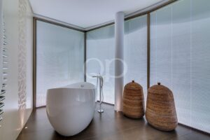 White freestanding oval bathtub in a modern bathroom with tall blinds and a light column, plus two woven wicker baskets by the window.
