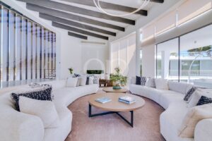 Bright modern living room with white curved sofas, two round coffee tables, beige rug, and large glass windows.