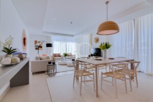 Bright open-plan living and dining area with white furniture, a wooden dining table, beige chairs, and large sheer-curtain windows.