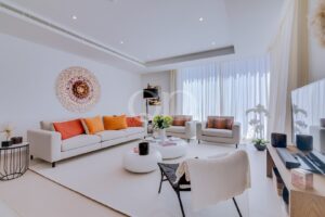 Contemporary white living room with beige sofas and orange cushions, a round wall sculpture, and sheer floor-to-ceiling windows.