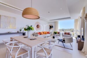Bright open-plan living and dining room with white furniture, a wooden dining table, and a large circular pendant lamp. Large windows show a sunny landscape outside.