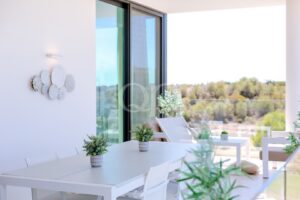 White outdoor dining area on a balcony with a long white table, matching chairs, and small potted plants; glass railing overlooks trees in the distance.