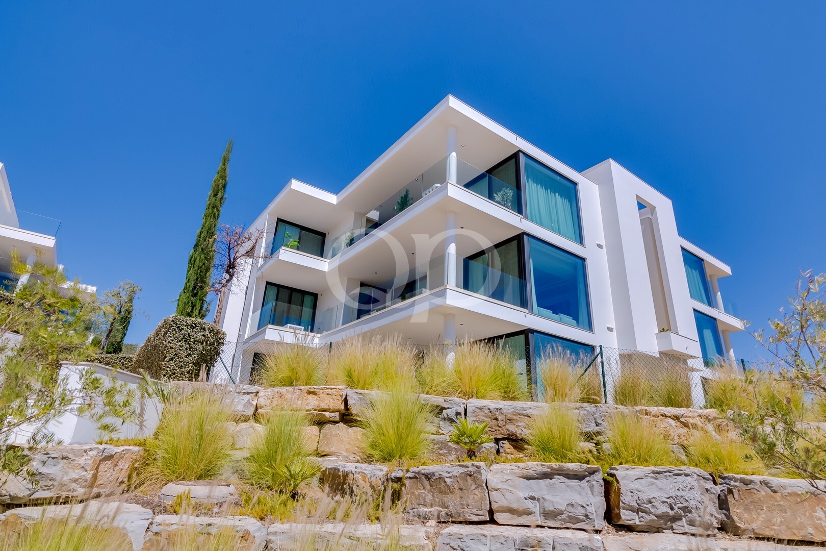 Modern white, multi-story house with large glass balconies on a rocky hillside under a clear blue sky.