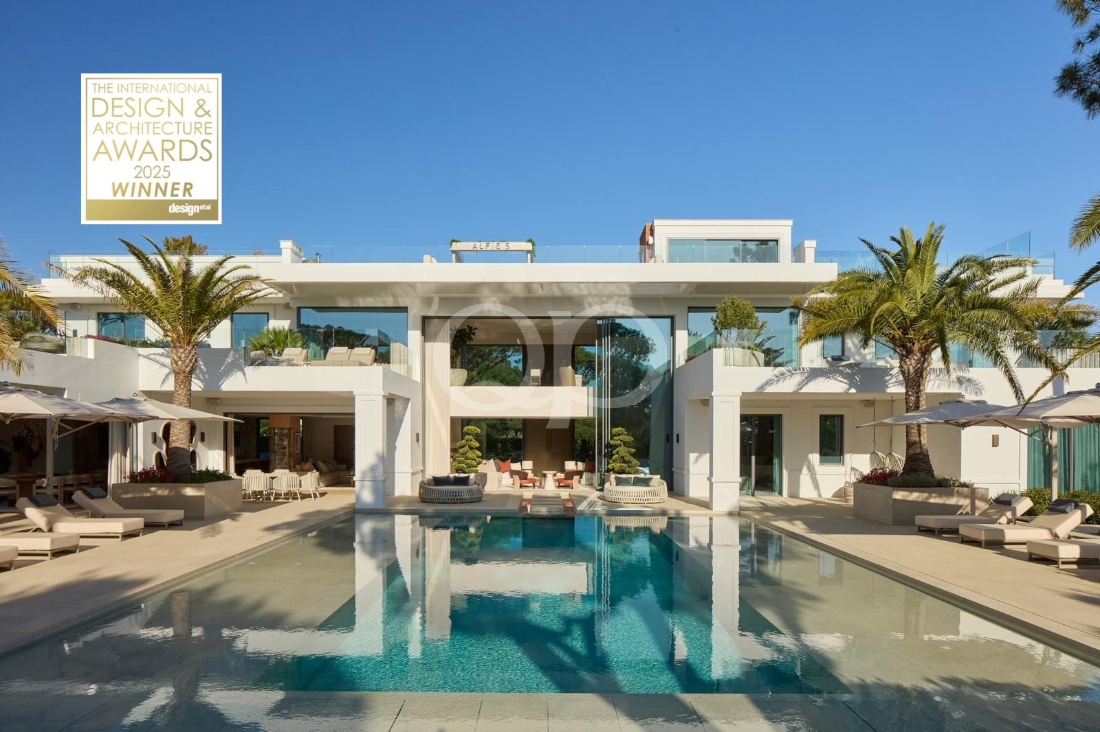 Modern white villa with a spacious pool deck, lounge chairs, and palm trees; award sticker in the upper left reads Design & Architecture Awards 2025 Winner.