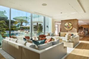 Bright open-plan living room with white sectional sofa, colorful cushions, and round coffee tables, overlooking a pool through floor-to-ceiling glass walls.