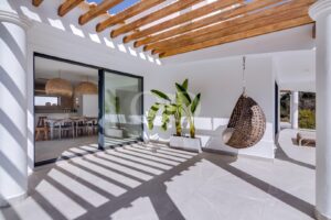 Pátio branco e ensolarado com uma pérgula de madeira, uma cadeira de vime suspensa, um vaso com uma planta e portas de vidro de correr que dão acesso a uma sala de jantar interior.
